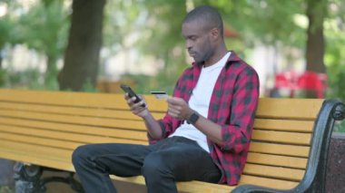 Başarılı çevrimiçi alışveriş için heyecanlı genç Afrikalı bir adam bir bankta otururken telefonla alışveriş yapıyor.