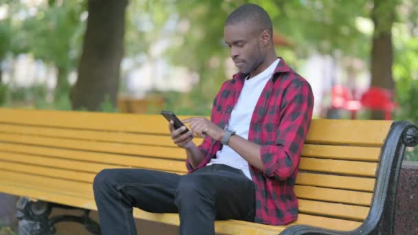 非洲年轻人坐在户外的长椅上 一边用智能手机庆祝成功 — 图库视频影像