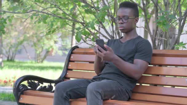 Молодой Африканский Человек Празднует Успех Смартфоне Сидя Скамейке Запасных — стоковое видео
