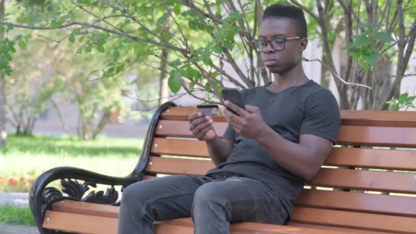 ベンチに座っている間に電話でオンラインショッピングをする若いアフリカ人男性 — ストック動画