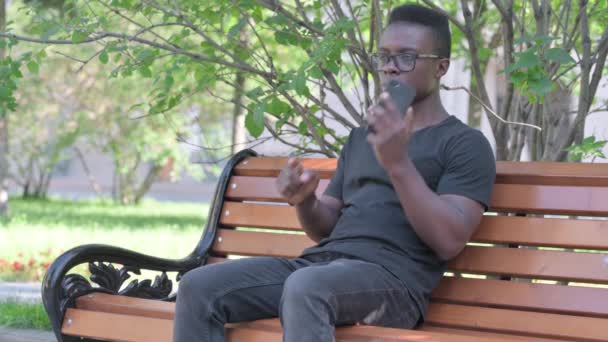 非洲年轻人坐在户外的长椅上 一边打电话一边聊天 — 图库视频影像