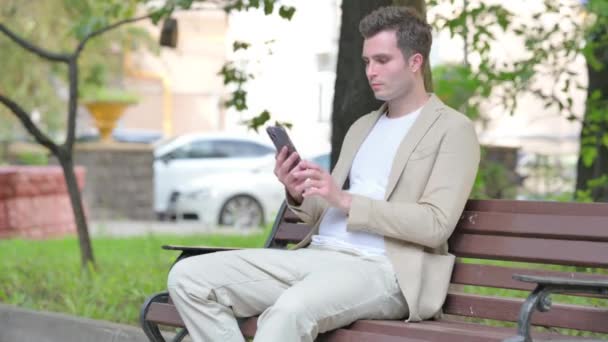 Nenucený Mladý Muž Slaví Úspěch Smartphone Zatímco Sedí Venku Lavičce — Stock video