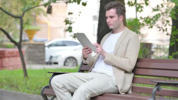 ベンチに屋外に座っている間 タブレットを使用しているカジュアルなヤングマン — ストック動画
