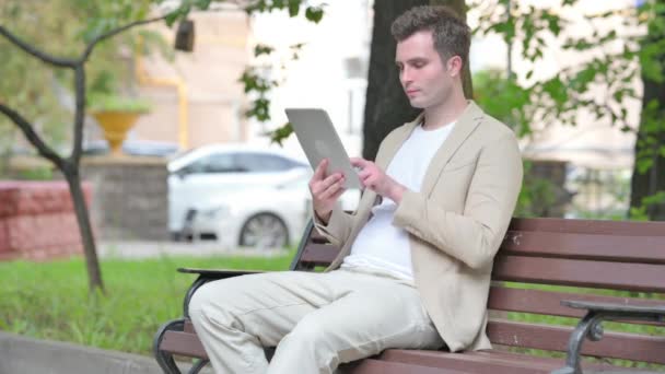 ベンチに屋外に座っている間 タブレット上の損失によってカジュアルな若い男が浮上します — ストック動画