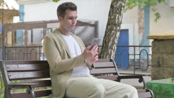 年轻人坐在户外的长椅上 一边用智能手机庆祝成功 — 图库视频影像