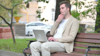 Sıradan genç adam dışarıda dizüstü bilgisayarda düşünüyor ve çalışıyor.