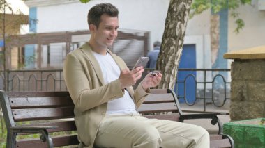 Genç adam dışarıda otururken telefonla alışveriş yapıyor.