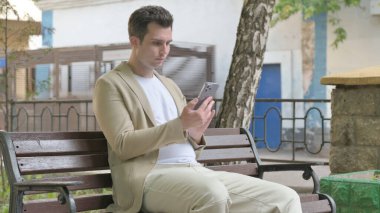 Genç adam dışarıda bankta otururken akıllı telefon kullanıyor.