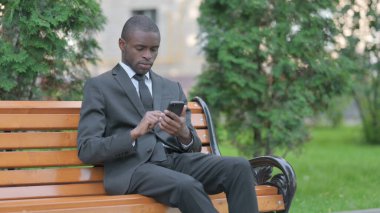 Bir bankta otururken akıllı telefon kullanan Afrikalı bir işadamı