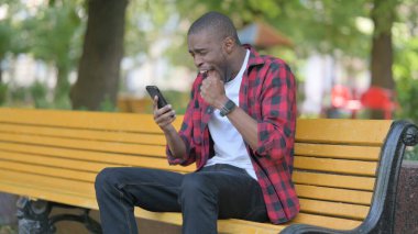 Genç Afrikalı Adam, bir bankta otururken akıllı telefonda başarıyı kutluyor.
