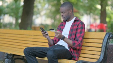 Başarılı çevrimiçi alışveriş için heyecanlı genç Afrikalı bir adam bir bankta otururken telefonla alışveriş yapıyor.