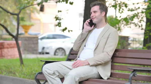 Casual Young Man Talking on phone while Sitting Outdoor on a Bench