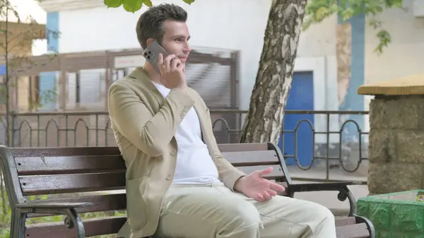 Young Man Talking on phone while Sitting Outdoor on a Bench