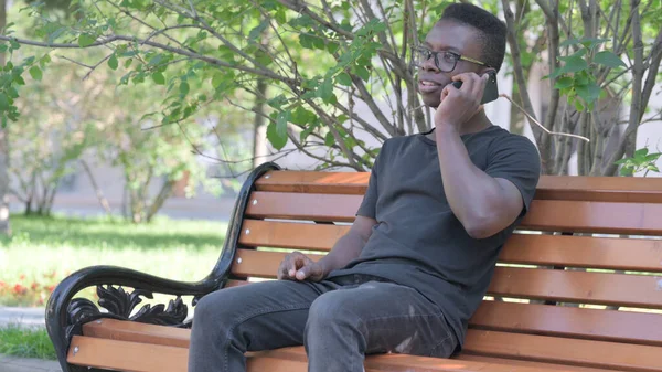 Young African Man Talking on phone while Sitting Outdoor on a Bench