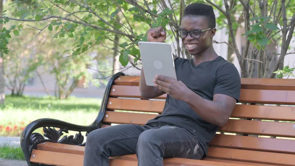 Young African Man Celebrating Success on Tablet while Sitting Outdoor on a Bench