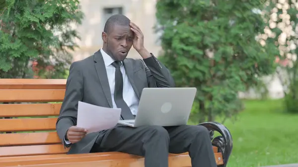African Businessman Feeling Upset while Reading Contract and Using Laptop Outdoor