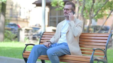 Orta yaşlı bir adam bankta otururken telefonla konuşuyor.