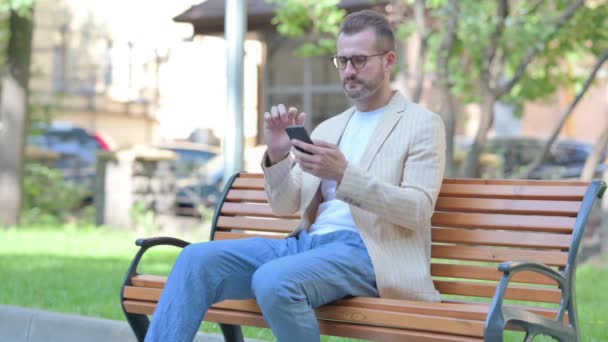 Středověký Muž Slaví Úspěch Smartphone Zatímco Sedí Venku Lavičce — Stock video