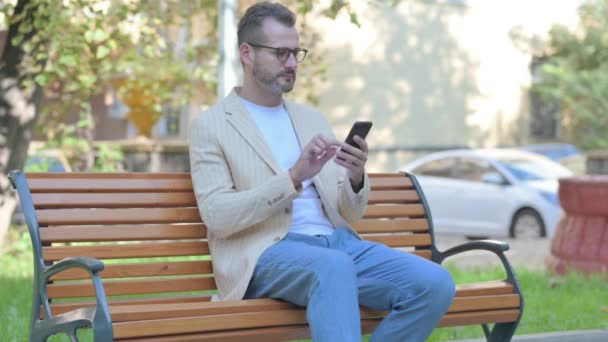 Nowoczesny Człowiek Casual Pomocą Smartfona Podczas Siedzenia Zewnątrz Ławce — Wideo stockowe