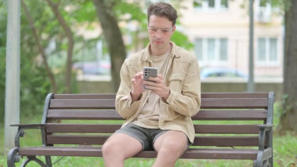 ベンチに屋外に座っている間 スマートフォンで損失にショックを受けたカジュアルな若者 — ストック動画