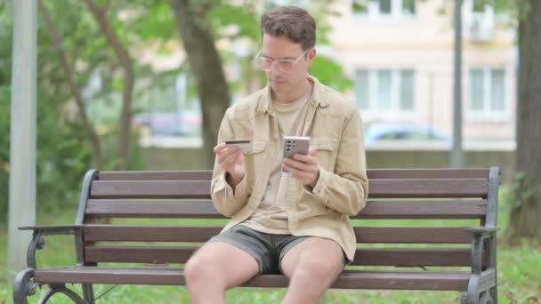 屋外に座っている間 電話でオンラインショッピングをするカジュアルなヤングマン — ストック動画