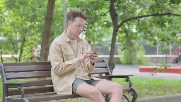 Nowoczesny Młody Człowiek Świętuje Smartfonie Siedząc Ławce — Wideo stockowe