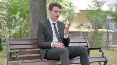 Genç İşadamı bir bankta otururken telefonda konuşuyor.