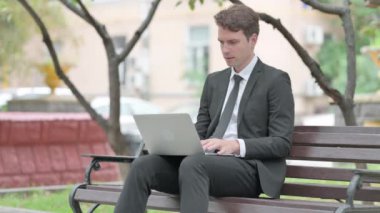 İş adamı kucağında laptopla kameraya bakıyor.