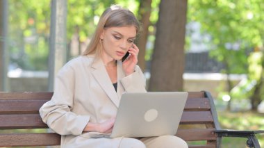 Genç iş kadını telefonda konuşuyor ve dışarıda dizüstü bilgisayar kullanıyor.