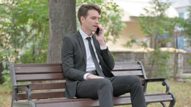 Genç İşadamı bir bankta otururken telefonda konuşuyor.