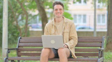Laptop üzerinde çalışırken kameraya gülümseyen sıradan bir genç adam.