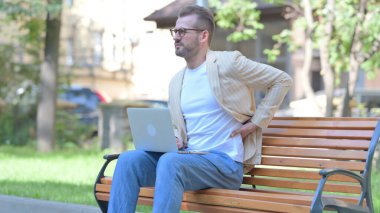 Orta yaşlı adam dışarıda otururken dizüstü bilgisayarla sırt ağrısı çekiyor