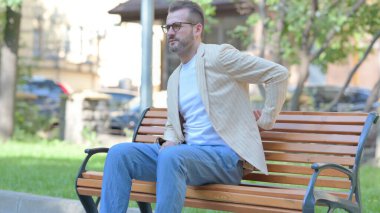 Orta yaşlı adam dışarıda sırt ağrısıyla oturuyor.