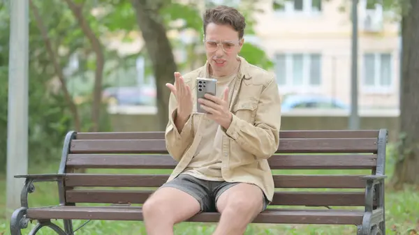 怒っているカジュアルな若い男は屋外に座っている間スマートフォンでビデオチャットを行います — ストック写真