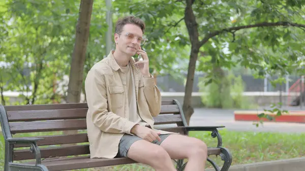 Modern Young Man Talking on phone while Sitting on Bench