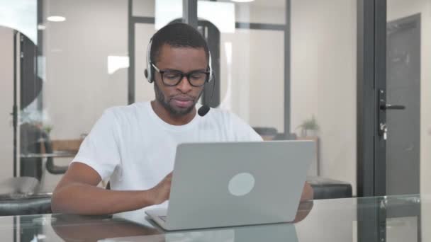 ラップトップで作業中にカメラを見ているヘッドセットを持つ若いアフリカ人男性 — ストック動画