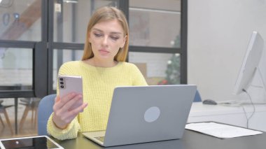 Laptop 'ta çalışan ve akıllı telefon kullanan genç sarışın kadın.