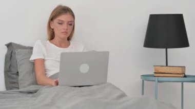 Sırt ağrısı olan genç bir kadın laptopta çalışırken yatakta oturuyor.