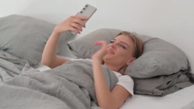 Yatakta uzanırken telefonda görüntülü konuşma yapan genç kadın.