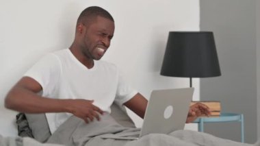 Sırt ağrısı olan Afrikalı Adam laptopta çalışırken yatakta oturuyor.