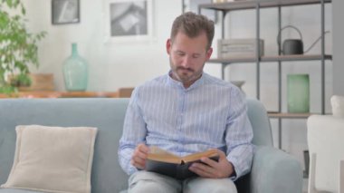 Orta yaşlı adam evdeki koltukta otururken kitap okuyor.