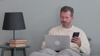 Yatakta dizüstü bilgisayarı olan orta yaşlı bir adam akıllı telefon kullanıyor.
