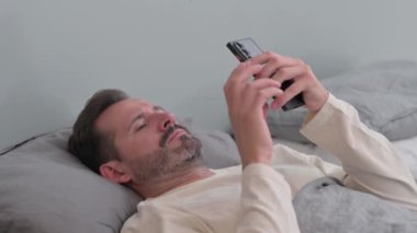 Orta yaşlı adam yatakta yatarken akıllı telefon kullanıyor.