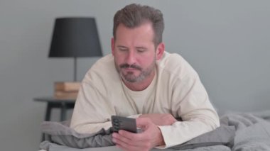 Adam yatakta karnı üzerinde yatarken telefonu kullanıyor.