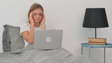 Başağrısı olan genç kadın laptopta çalışırken yatakta oturuyor.