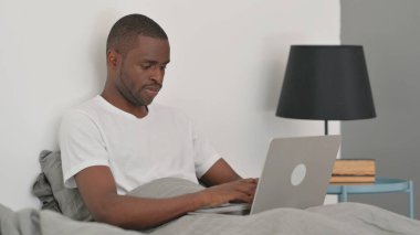 Yatakta otururken dizüstü bilgisayarda çalışan Afrikalı Adam