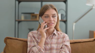 Evdeki kanepede oturan genç bir kadın telefonda konuşuyor.