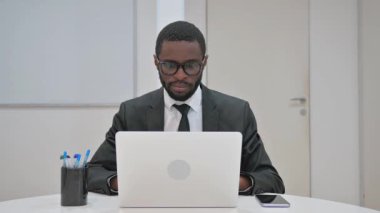 Hevesli Afro-Amerikan iş adamı, ofis masasındaki kameraya dizüstü bilgisayarla selam veriyor.