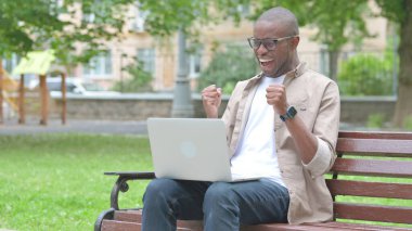 Dizüstü bilgisayarlı neşeli genç Afrikalı Park Bench 'te kazanmanın tadını çıkarıyor.