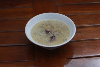 a close up of a bowl of green bean porridge and black sticky rice doused in coconut milk, served warm and ready to eat on a wooden background with a natural pattern. Indonesian food concept.